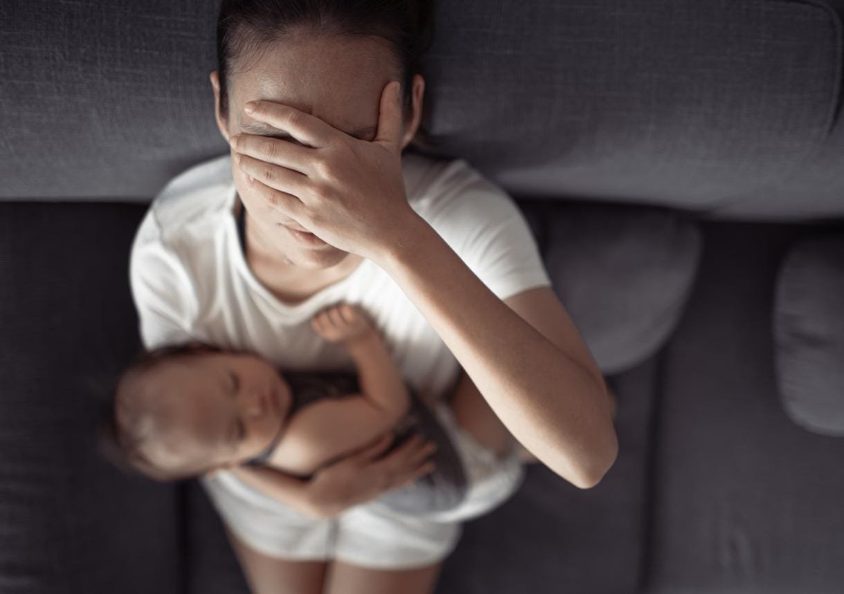 COPY_istock-1420207647-1698056736 Une mère levant la tête tout en se couvrant les yeux et tenant son bébé, pour manifester sa dépression.