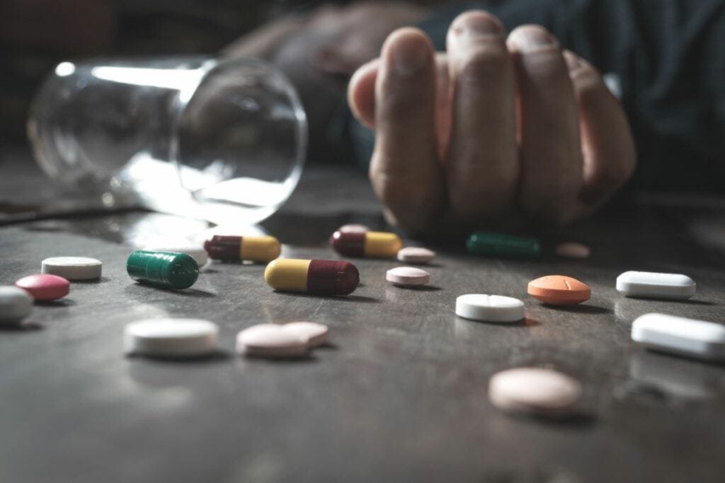 Drug-Overdose-Blog-1200-1024×683 Un victime d'overdose étendu au sol avec plein de médicament éparpillé autour.