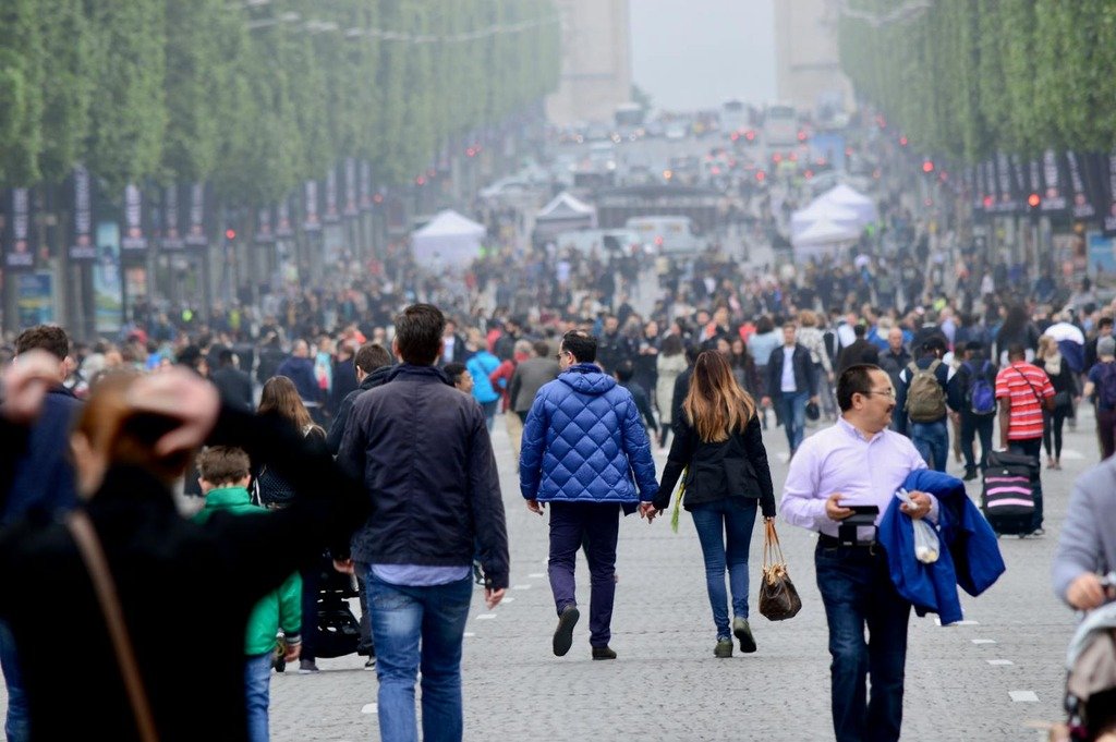 A vibrant crowd walking on the famous Champs-Élysées in Paris, with the Arc de Triomphe in the background.
