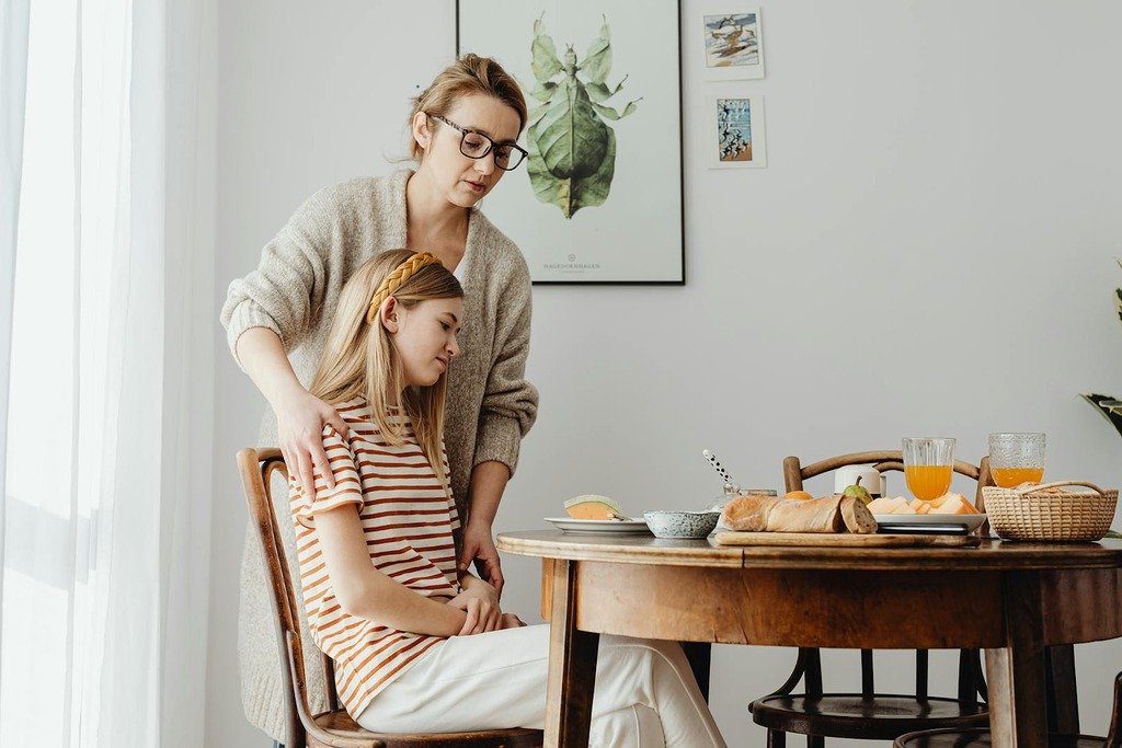 A mother comforts her teenage daughter during breakfast. A warm family moment at the dining table.