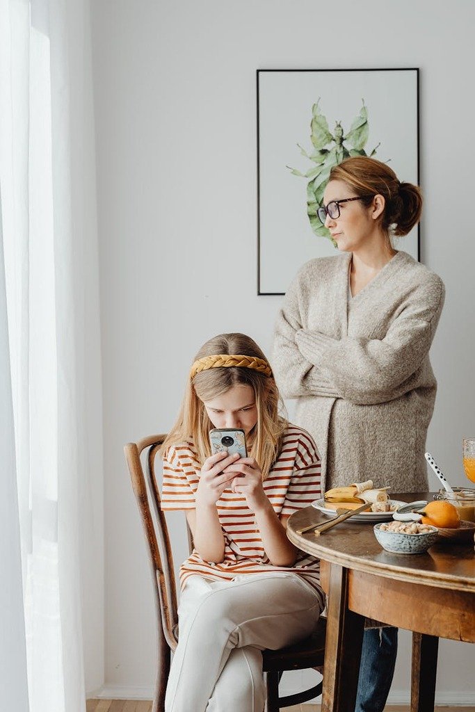 A mother with crossed arms stands beside a daughter using a smartphone at breakfast.