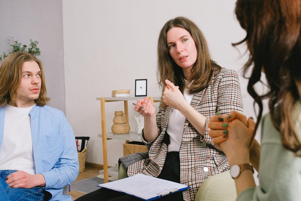 Thoughtful woman psychologist in stylish clothes siting with clipboard and talking with couple in light room