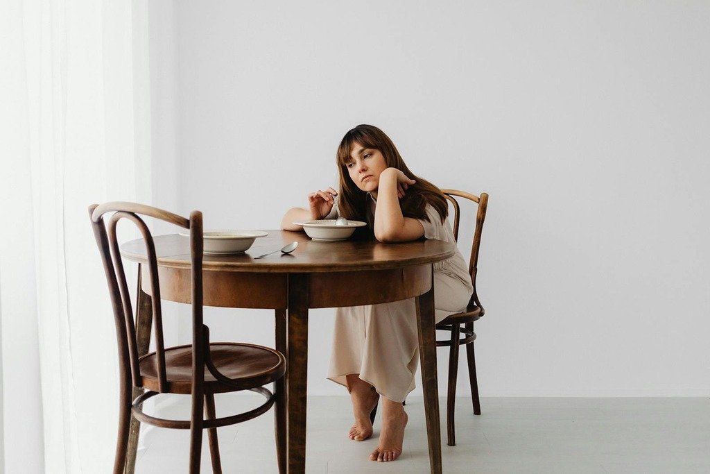 A woman sits pensively at a wooden table with a bowl, conveying a mood of contemplation.