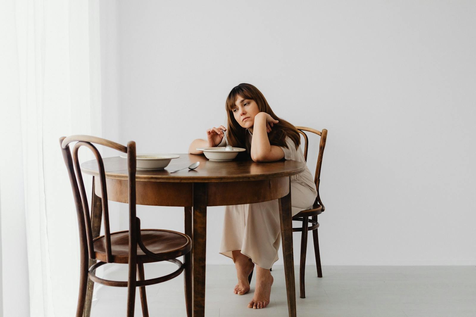 A woman sits pensively at a wooden table with a bowl, conveying a mood of contemplation.