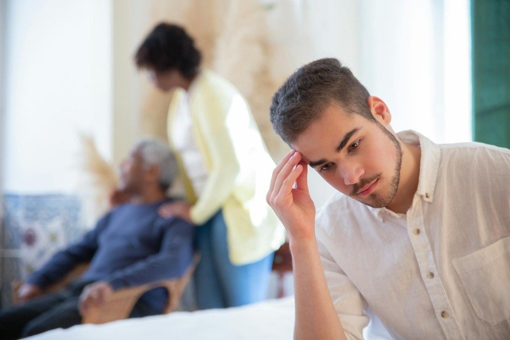 Young man in thoughtful pose indoors with family members in the background, expressing contemplation and care.