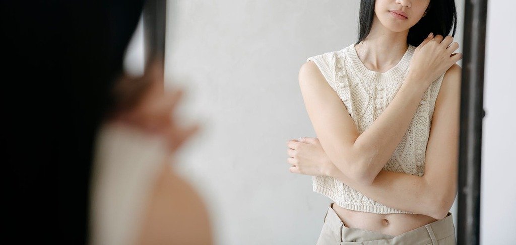 Reflection of crop young female in sleeveless shirt gently embracing herself while looking at mirror