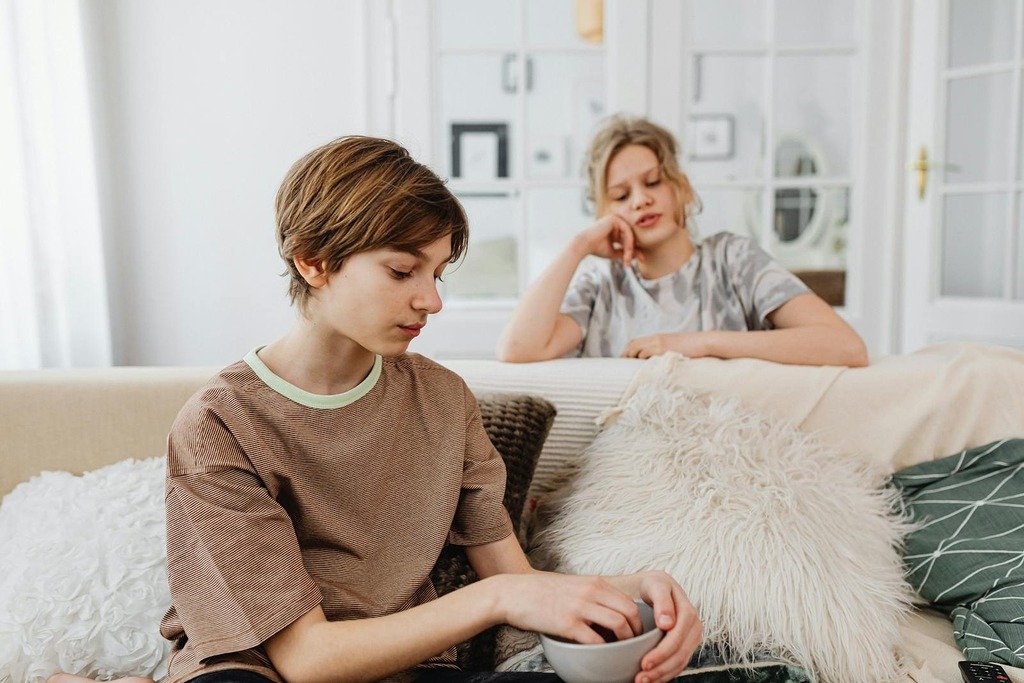 Two teenagers sitting on a comfortable sofa, one holding a bowl, in a well-lit living room.