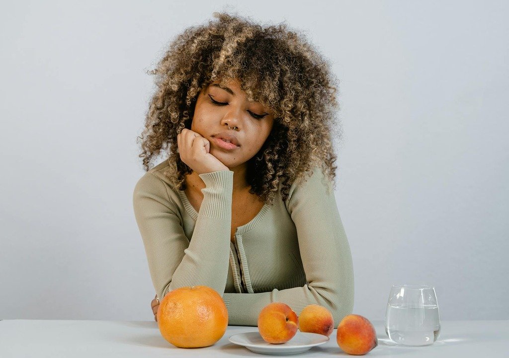 A woman with curly hair in green sleeves sits with oranges and peaches on a white table.
